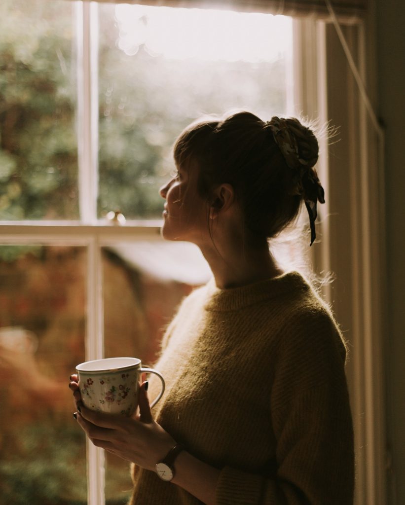 Young woman holding a floral mug and gazing out of a window in soft morning light, reflecting on the quiet moments that come with living where you work as a live-in carer.
