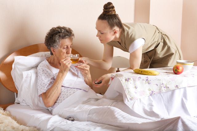 A caregiver demonstrating duty of care by assisting an elderly woman in bed with drinking tea. The caregiver is attentively supporting the woman while a tray with fruit and a bowl is placed nearby, reflecting a nurturing and safe environment.