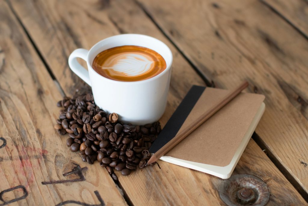 A cup of coffee with latte art sits on a rustic wooden table beside scattered coffee beans, a notebook, and a pencil — symbolising taking break in the live in care.